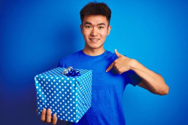 Asyalı genç bir Çinli doğum günü hediyesi tutuyor. İzole edilmiş mavi arka planda duruyor. Yüzü kendini işaret ediyor.