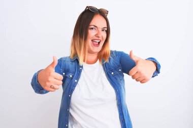Güzel bir kadın kot gömlek giyer, izole edilmiş beyaz arka planda durur. Elle olumlu bir jest yapar, gülümser ve başarı için mutlu olur. Kazanan jest.