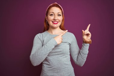 Gri tişört giyen güzel, kızıl saçlı bir kadın ve mor arka planda izole edilmiş bir çember var. Gülümsüyor ve kameraya bakıyor. İki eliyle ve parmaklarıyla yüzünü gösteriyor..