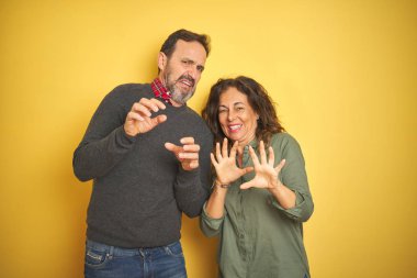 Güzel bir orta yaşlı çift, izole edilmiş sarı arka planda tiksinti ifadesi, hoşnutsuz ve tiksindirici bir ifade, tiksinti reaksiyonu yüzünden tiksinti duyuyorlar. Eller havada. Can sıkıcı bir kavram..