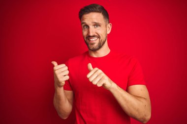 Kırmızı izole edilmiş arka plan üzerine gündelik tişört giyen genç yakışıklı adam el ve baş parmaklarıyla arka tarafı işaret ediyor, kendinden emin gülümsüyor.