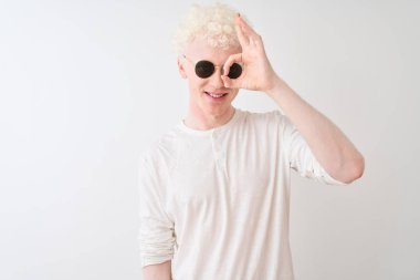 Genç albino tişört ve güneş gözlüğü takan adam izole edilmiş beyaz arka planda el hareketleriyle gülümsüyor ve mutlu yüzle parmaklarının arasından bakıyor..