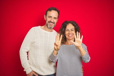 Güzel orta yaşlı çift, izole edilmiş kırmızı arka plan üzerine kışlık süveter giyip 8 numaralı parmaklarıyla işaret ederken kendinden emin ve mutlu bir şekilde gülümsüyor..