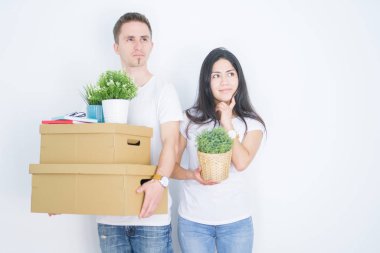 Young beautiful couple holding cardboard boxes standing over isolated white background serious face thinking about question, very confused idea