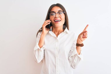 Gözlüklü genç iş kadını izole edilmiş beyaz arka planda akıllı telefondan konuşuyor. Elleri ve parmakları kenarı işaret ederek çok mutlu.