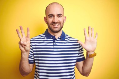 Sakallı genç ve kel bir adam sarı izole edilmiş arka plan üzerine günlük mavi tişört giyiyor ve 8 numarayı gösterirken kendinden emin ve mutlu bir şekilde gülümsüyor..