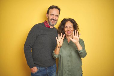 Güzel orta yaşlı çift, izole edilmiş sarı arka planda dokuz numaralı parmaklarını gösterip gülümserken mutlu ve kendinden emin..