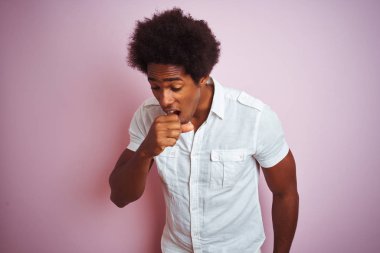 Afro saçlı, beyaz tişörtlü, izole edilmiş pembe arka planda duran genç Amerikalı adam kendini iyi hissetmiyor ve soğuk algınlığı veya bronşitin belirtisi olarak öksürüyor. Sağlık hizmetleri kavramı.