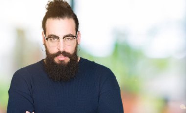Uzun saç ve sakal yüz çapraz kolları üzerinde gözlük şüpheci ve sinir, onaylamayan ifade giyen genç hipster adam. Negatif kişi.