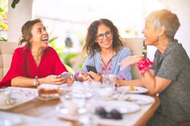 Orta yaşlı kadınlarla buluşup, öğle yemeği yiyip, kahve içiyorlar. Olgun arkadaşlar mutlu bir şekilde gülümsüyor. Güneşli bir günde evde akıllı telefon kullanıyorlar.
