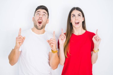 Genç ve güzel bir çift birlikte beyaz bir zemin üzerinde hayret ve şaşkınlık içinde yukarı bakıyor ve parmaklarıyla işaret ediyor ve kollarını kaldırıyor..