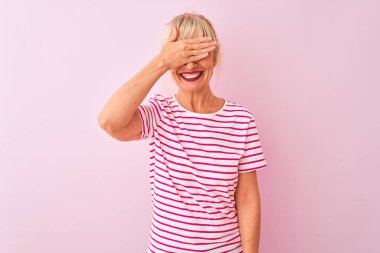 İzole edilmiş pembe arka planda duran çizgili tişört giyen orta yaşlı bir kadın. Gülümsüyor ve elleri yüzünde şaşkınlıktan gözleri kapatıyordu. Kör kavram.