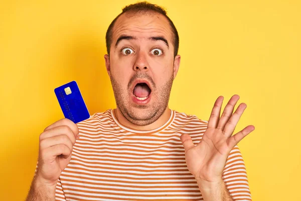 Молодой Человек Держащий Синюю Кредитку Стоящую Изолированном Желтом Фоне Счастлив — стоковое фото