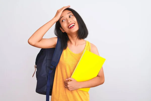 Young chinese student woman wearing backpack holding book over isolated white background stressed with hand on head, shocked with shame and surprise face, angry and frustrated. Fear and upset for mistake.