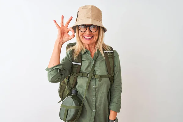 Middle age hiker woman wearing backpack hat canteen glasses over isolated white background smiling positive doing ok sign with hand and fingers. Successful expression.