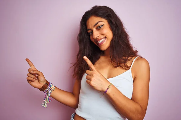Young beautiful woman wearing white t-shirt standing over isolated pink background smiling and looking at the camera pointing with two hands and fingers to the side.