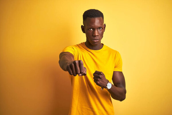 Young african american man wearing casual t-shirt standing over isolated yellow background Punching fist to fight, aggressive and angry attack, threat and violence