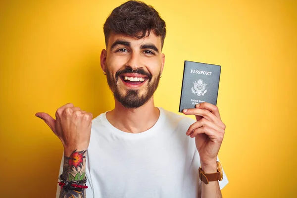 Young man with tattoo wearing United States USA passport over isolated yellow background pointing and showing with thumb up to the side with happy face smiling
