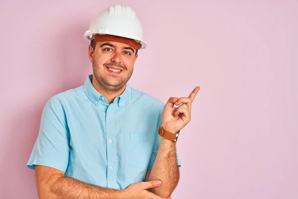 年轻的建筑师戴着安全帽 站在孤立的粉色背景上 脸上挂着大大的笑容 手指手画脚地看着摄像机的侧面 — 图库照片