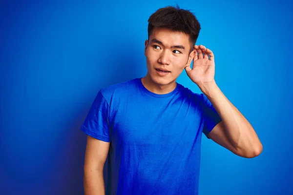 Tシャツを着た若いアジア系中国人男性は 噂やゴシップに耳を傾けながら 手で笑みを浮かべて孤立した青い背景の上に立っています 病気の概念 — ストック写真