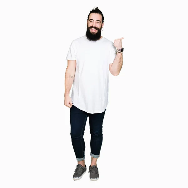 年轻的时髦男子长头发和胡须穿着休闲的白色T恤微笑与快乐的脸看 并指着一边竖起大拇指 — 图库照片