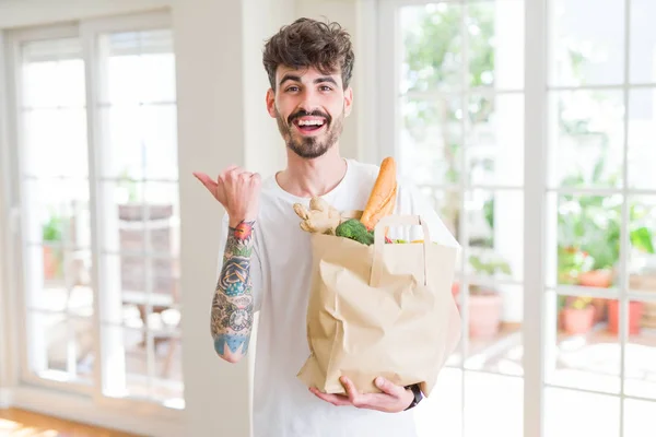 Young man holding paper bag of fresh groceries from the supermarket pointing and showing with thumb up to the side with happy face smiling