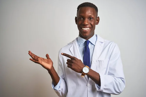Young african american doctor man wearing coat standing over isolated white background amazed and smiling to the camera while presenting with hand and pointing with finger.