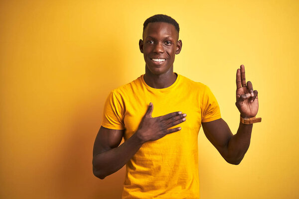 Young african american man wearing casual t-shirt standing over isolated yellow background smiling swearing with hand on chest and fingers up, making a loyalty promise oath