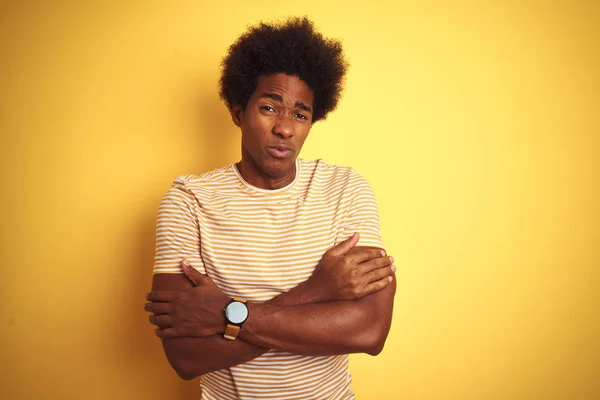 American man with afro hair wearing striped t-shirt standing over isolated yellow background shaking and freezing for winter cold with sad and shock expression on face
