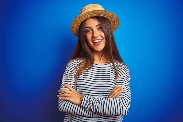 Young beautiful woman wearing navy striped t-shirt and hat over isolated blue background happy face smiling with crossed arms looking at the camera. Positive person.
