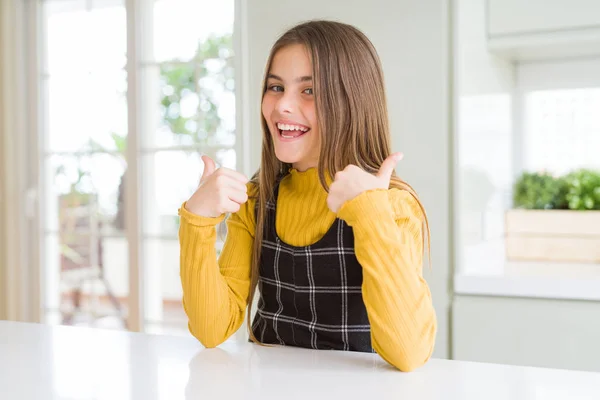 Young beautiful blonde kid girl wearing casual yellow sweater at home success sign doing positive gesture with hand, thumbs up smiling and happy. Cheerful expression and winner gesture.