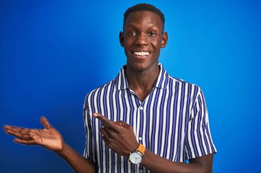 İzole edilmiş mavi arka plan üzerinde duran çizgili günlük gömleği giyen Afrikalı Amerikalı adam kameraya hayret ediyor ve gülümsüyor..