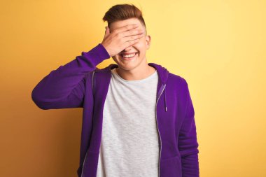 Mor eşofman üstü giyen genç yakışıklı adam izole edilmiş sarı arka planda duruyor. Gülümsüyor ve elleri yüzünde şaşkınlıktan gözleri kapatıyordu. Kör kavram.