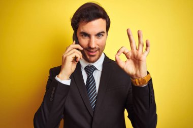 Akıllı telefondan konuşan genç yakışıklı iş adamı izole edilmiş sarı arka plan üzerinde tamam işareti yapıyor, mükemmel bir sembol.