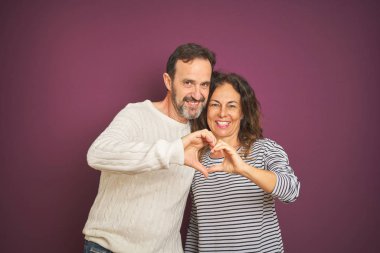 Güzel bir orta yaşlı çift, izole edilmiş mor arka plan üzerine kazak giyip birbirlerine gülümsüyorlar. Elleriyle kalp sembolü yapıyorlar. Romantik konsept.