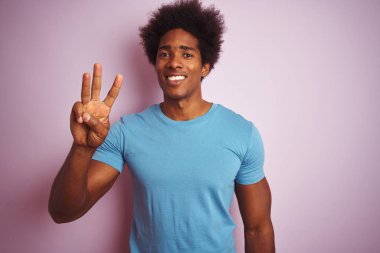 Afro saçlı, mavi tişörtlü, izole edilmiş pembe arka planda duran Afro-Amerikalı adam üç numarayı gösterirken kendinden emin ve mutlu bir şekilde gülümsüyor..