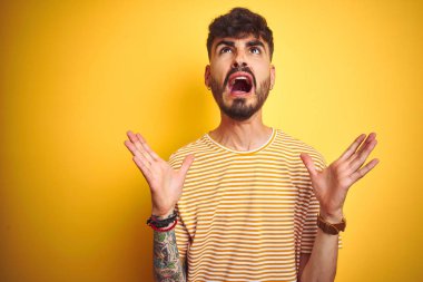 Dövmeli genç adam çizgili t-shirt giyen izole sarı arka plan üzerinde duran deli ve deli bağırarak ve agresif ifade ve kollar kaldırdı ile bağırıyor. Hayal kırıklığı kavramı.