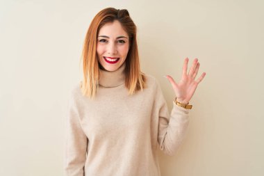 Kırmızı saçlı kadın zarif balıkçı yaka kazak giyiyor. İzole edilmiş beyaz arka plan üzerinde duruyor. Beş numarayı işaret ediyor ve gülümsüyor..