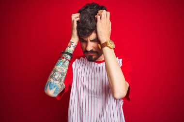 Çizgili tişörtlü genç adam izole edilmiş kırmızı arka planda dikiliyor. Ağrı ve migren yüzünden baş ağrısı çekiyor ve stresli. Ellerini başının üstüne koy..