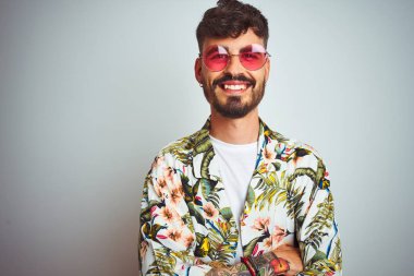 Tatilde dövmeli, güneş gözlüğü takan, izole edilmiş beyaz arka planda mutlu yüzü olan, kollarını kavuşturup kameraya bakan bir adam. Pozitif kişi.