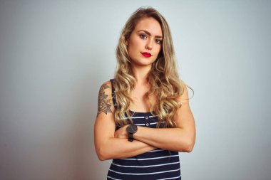 İzole edilmiş beyaz arka plan üzerinde duran çizgili tişört giyen genç ve güzel bir kadın şüpheci ve gergin, yüzünde çapraz kollu tasvip etmeyen bir ifade. Negatif kişi.