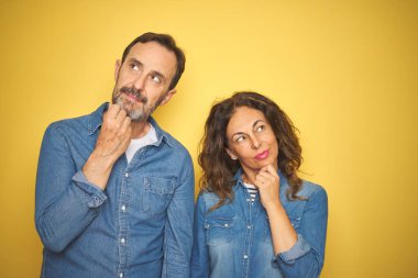 Güzel orta yaş çift birlikte izole sarı arka plan üzerinde el çene hakkında soru, dalgın ifade düşünme üzerinde ayakta. Düşünceli bir yüz ile gülümsüyor. Şüphe kavramı.