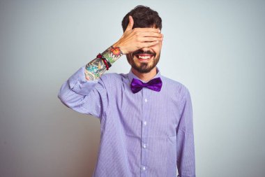 Mor gömlekli ve papyonlu genç adam izole edilmiş beyaz arka planda gülümsüyor ve elleri yüzünde şaşkınlıktan gözleri kapayarak gülüyor. Kör kavram.