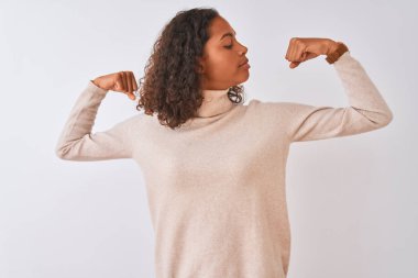 Balıkçı yaka kazak giyen genç Brezilyalı kadın izole beyaz arka planda dikiliyor ve gururla gülümseyen kol kaslarını gösteriyor. Fitness konsepti.