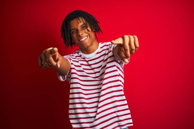 Rastalı tişörtlü genç afro adam izole edilmiş kırmızı arka planda dikiliyor. Sana ve kameraya parmaklarıyla bakıyor, neşeli ve pozitif gülümsüyor.