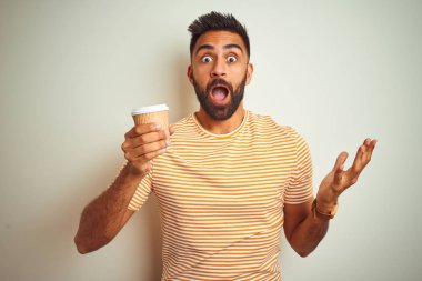 Genç Hintli bir adam izole edilmiş beyaz arka planda dikilmiş kahve içiyor. Çok mutlu ve heyecanlı. Kazanan ifade zaferi kutluyor.
