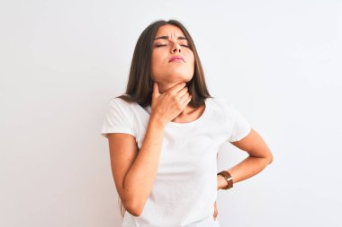 İzole edilmiş beyaz arka plan üzerinde duran sıradan tişört giyen genç ve güzel bir kadın ağrıyan boynuna dokunuyor, grip, mantar ve enfeksiyon için boğaz ağrısı çekiyor.