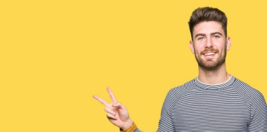 Çizgili kazak giyen genç yakışıklı adam mutlu yüz le gülümseyerek zafer işareti yaparak kameraya göz kırpıyor. İki numara.