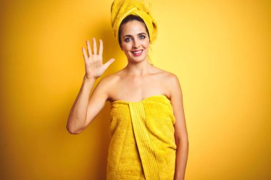 Güzel bir kadın duş havlusunu vücuduna ve başına takmış sarı izole arka planda beş numaralı parmaklarıyla işaret ederken kendinden emin ve mutlu bir şekilde gülümsüyor..