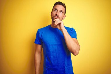 Sarı izole arka plan üzerine sıradan mavi tişört giyen genç yakışıklı bir adam. Elleri çenesinde soru sorarken, düşünceli bir ifade takınıyor. Düşünceli bir yüzle gülümsüyor. Şüphe kavramı.